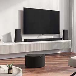 Modern living room with a wall-mounted flat-screen TV, two black speakers, a white floating console, and a round black coffee table on wooden flooring. Cozy atmosphere.