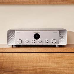 Silver audio amplifier with a sleek, modern design featuring circular knobs, central display, and metallic finishes, placed on a wooden shelf.