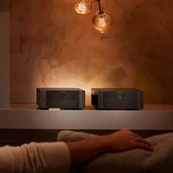 Dimly lit room with two small black devices on a shelf. A person’s arm rests on a couch below. Warm light from bulbs creates a cozy atmosphere.