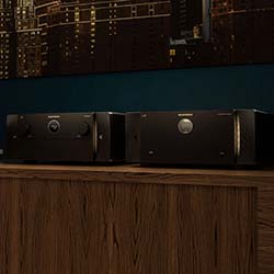 Two black audio amplifiers sit on a wooden table against a cityscape backdrop in dim lighting, creating a sleek and modern ambiance.