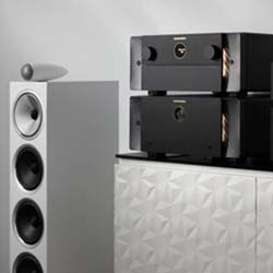 A sleek modern audio setup with a tall, white speaker featuring multiple black drivers next to two stacked black amplifiers on a textured cabinet.
