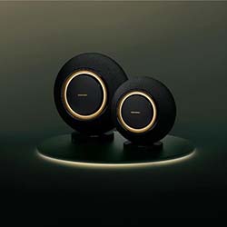 Two black round speakers with gold accents are placed on a glowing platform against a dark background, conveying a sleek, modern design.