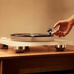 A hand lowers the needle onto a sleek, modern turntable on a wooden table. The warm lighting creates a cozy, nostalgic atmosphere.