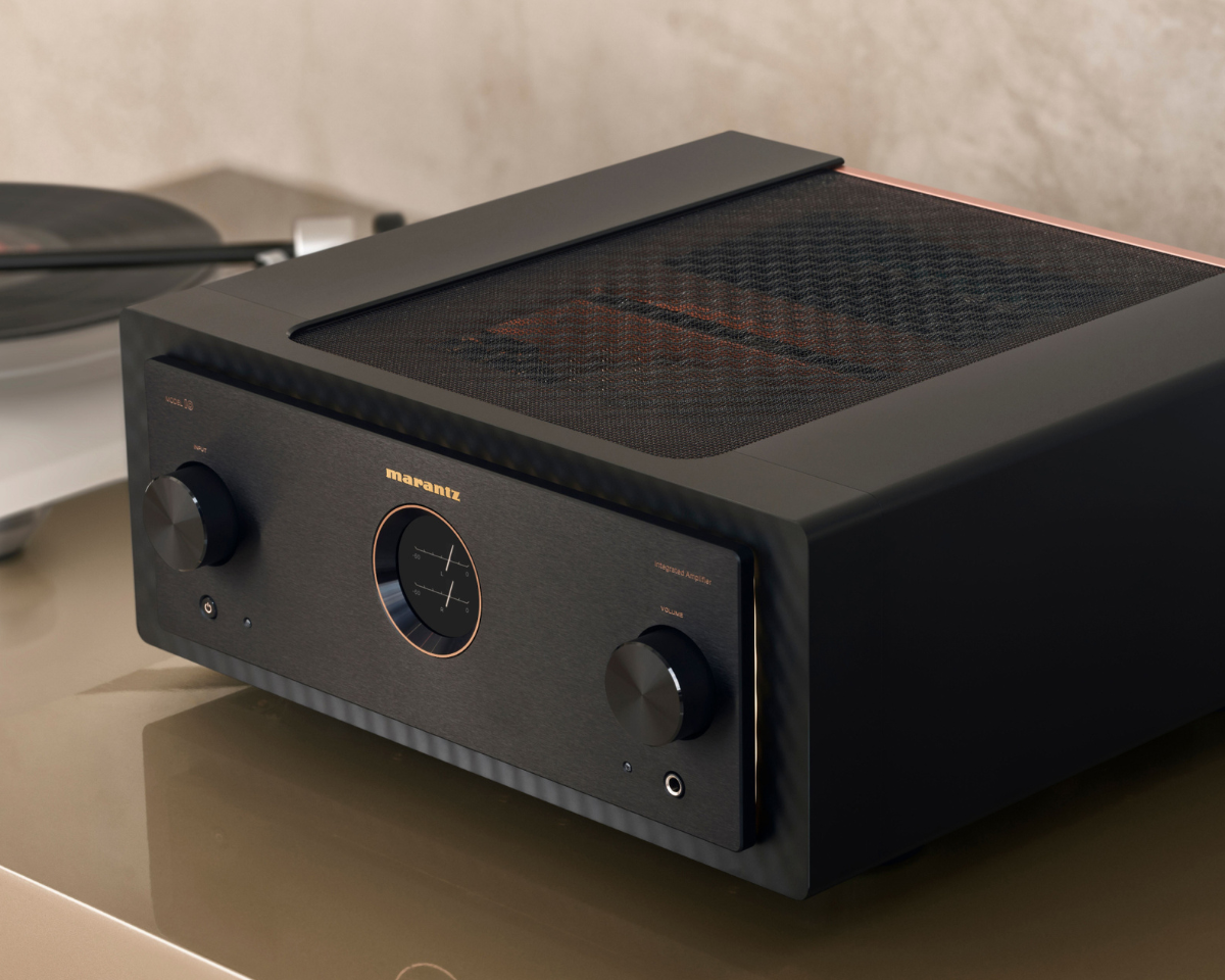 A sleek black audio amplifier with copper accents on a beige surface. Large knobs on the front, with a vinyl record player in soft focus behind it. Modern and elegant.