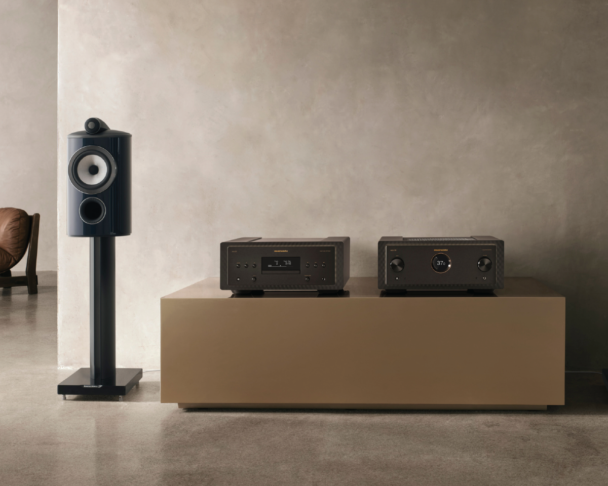 A sleek audio setup features a modern black speaker on a stand next to a beige table with two black electronic devices, set against a minimalist beige wall.