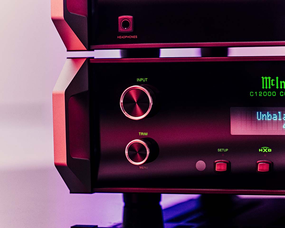Close-up of a sleek audio amplifier with illuminated knobs labeled "Input" and "Trim." A digital display reads "Unbalanced." Pink and purple lighting creates a modern, sophisticated tone.