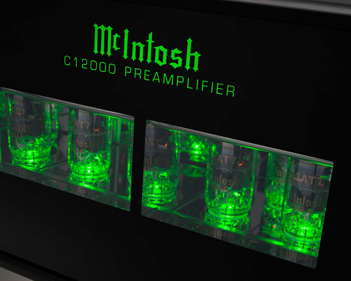 Close-up of a McIntosh C12000 preamplifier. Bright green glow from six vacuum tubes behind a glass panel contrasts with the glossy black surface.