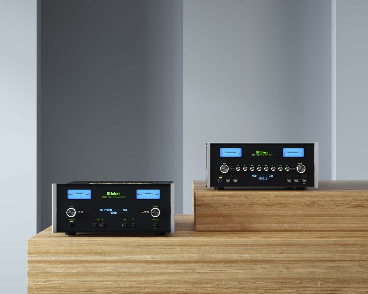 Two high-end audio amplifiers with glowing blue screens and silver knobs are displayed on wooden platforms against a minimalist gray wall.