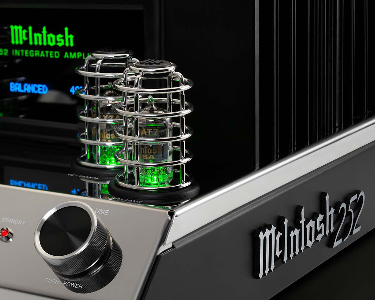 Close-up of a McIntosh amplifier with glowing green tubes and a sleek metal finish. The front panel features a volume knob and backlit logo.