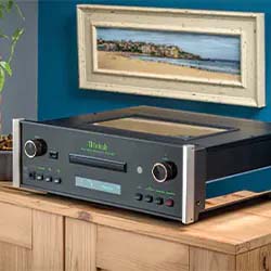 A vintage stereo amplifier with a wood panel sits on a wooden table against a blue wall. Above it hangs a framed painting of a seaside landscape.