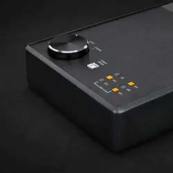 A black electronic audio interface with a large round knob and several colorful buttons is pictured against a dark background, conveying a sleek, modern tone.