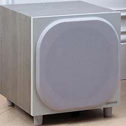 A light gray subwoofer with a cube shape and rounded edges on the front panel sits on a wooden floor. The design is sleek and modern, giving a minimalist vibe.