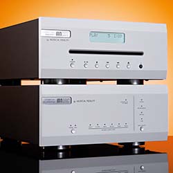Two stacked silver audio components against an orange background. The top unit shows a display reading "PLAY". Reflective black surface below.