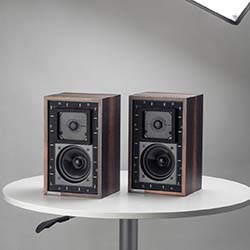 Two vintage-style speakers with wooden casing and black fronts are placed side by side on a round white table, against a neutral gray background.