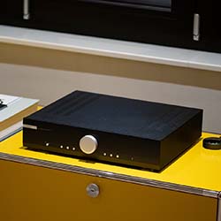 A sleek black audio amplifier with a large silver dial sits on a bright yellow cabinet in a dimly lit room, conveying modern and elegant design.