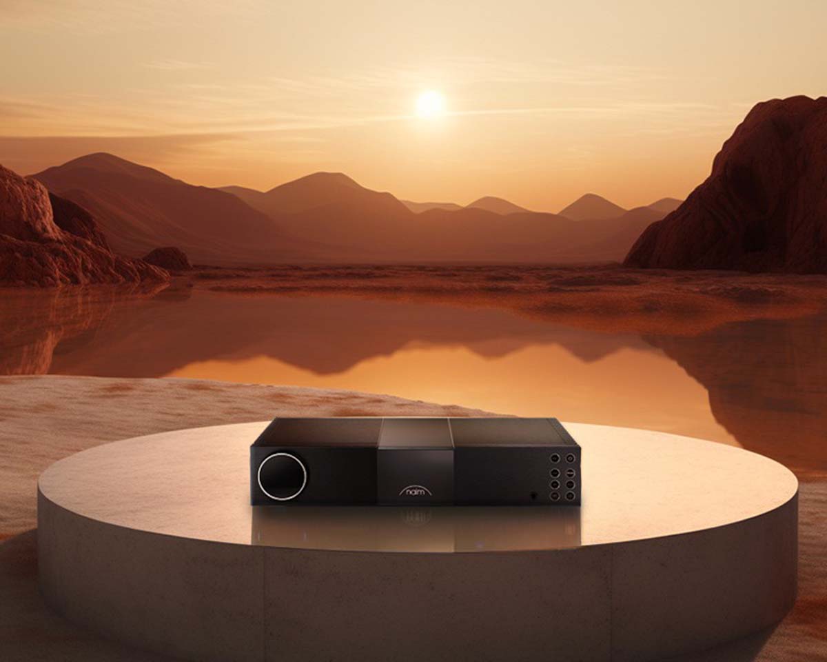 A sleek black audio device sits on a round, white platform against a serene landscape of a desert at sunset, with orange skies and distant mountains.