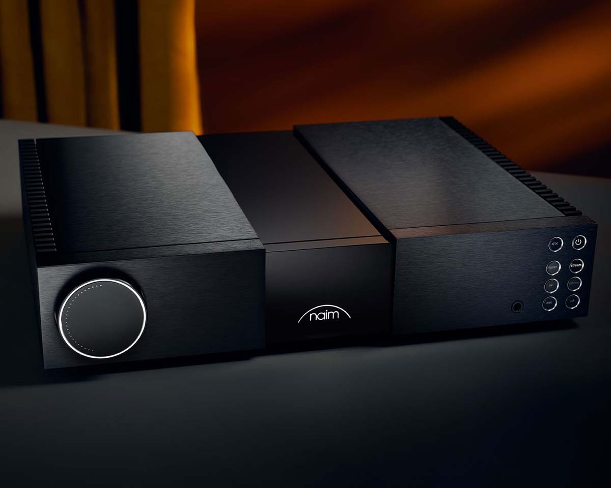 Sleek black audio amplifier with a large volume dial on the left, circular buttons on the right, and the "naim" logo at the center, set against a warm backdrop.