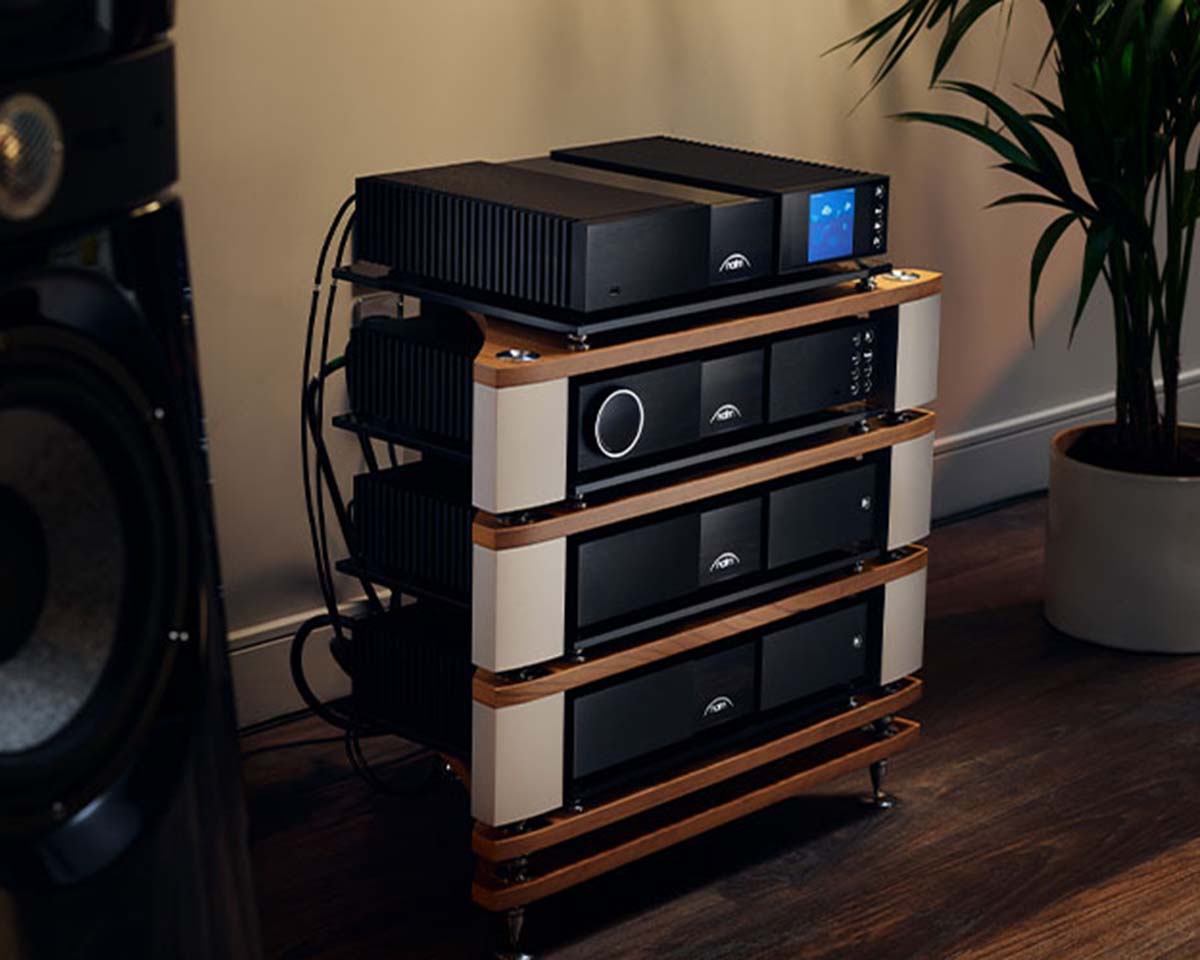 A sleek audio equipment setup on a wooden rack with multiple black units, buttons, and a display screen. It's in a cozy room with warm lighting and a potted plant nearby.