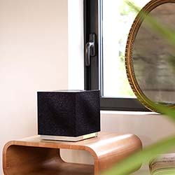 A black cube speaker sits on a wooden table by a window. The scene is minimalist with soft lighting, conveying a calm and modern atmosphere.