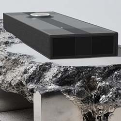 A sleek, black rectangular speaker sits on a rugged, metallic textured surface. The modern design contrasts with the organic, rocky texture below.