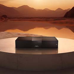 A sleek black electronic device sits on a circular platform against a tranquil desert landscape at sunset, conveying a sense of calm and modern elegance.