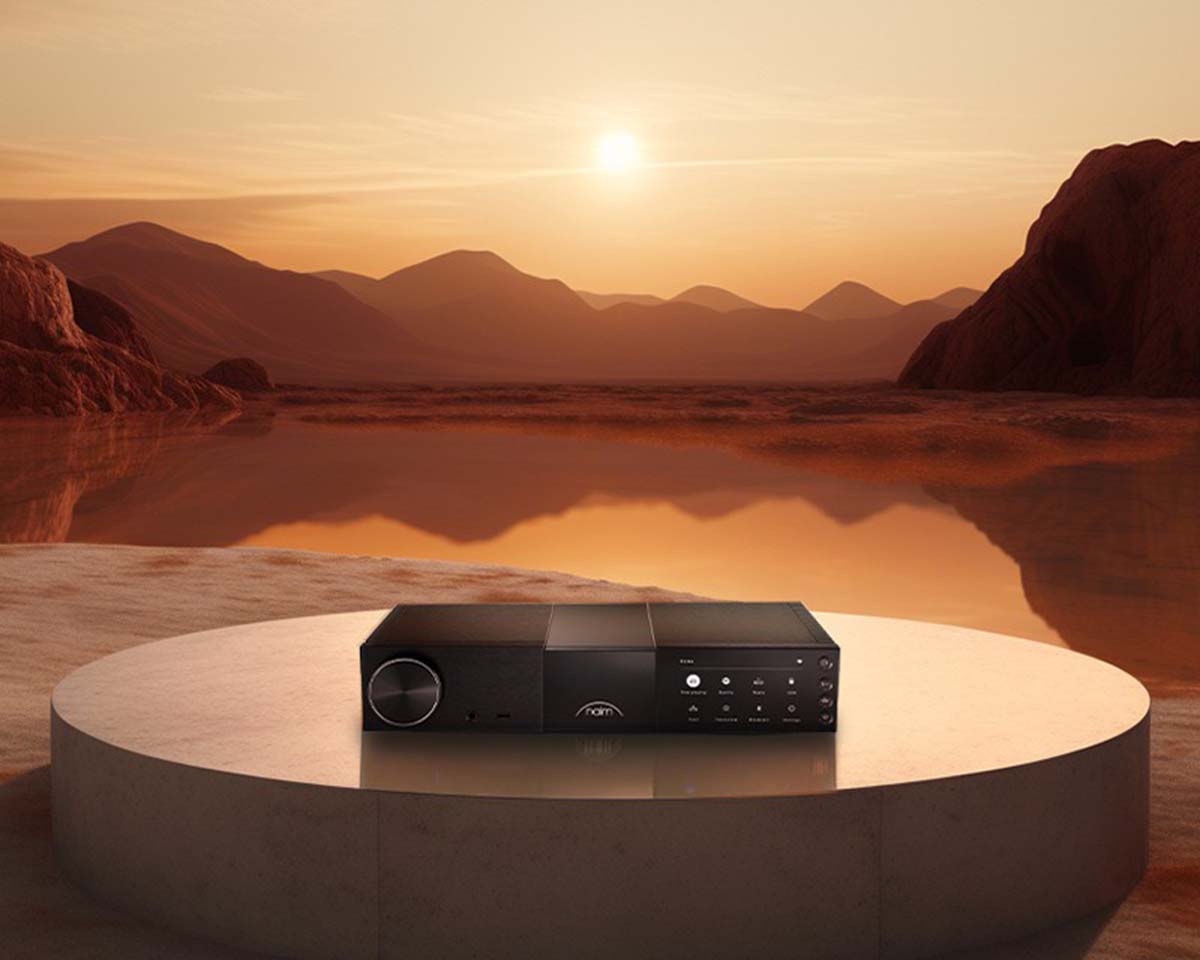 A sleek audio system rests on a circular platform against a serene desert backdrop at sunset, with warm hues reflecting off a calm water surface.