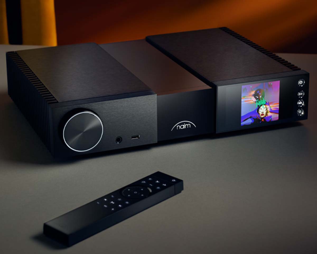 Naim audio device with a sleek black design on a table, featuring a large knob, touchscreen display showing album art, and a remote control in the foreground.