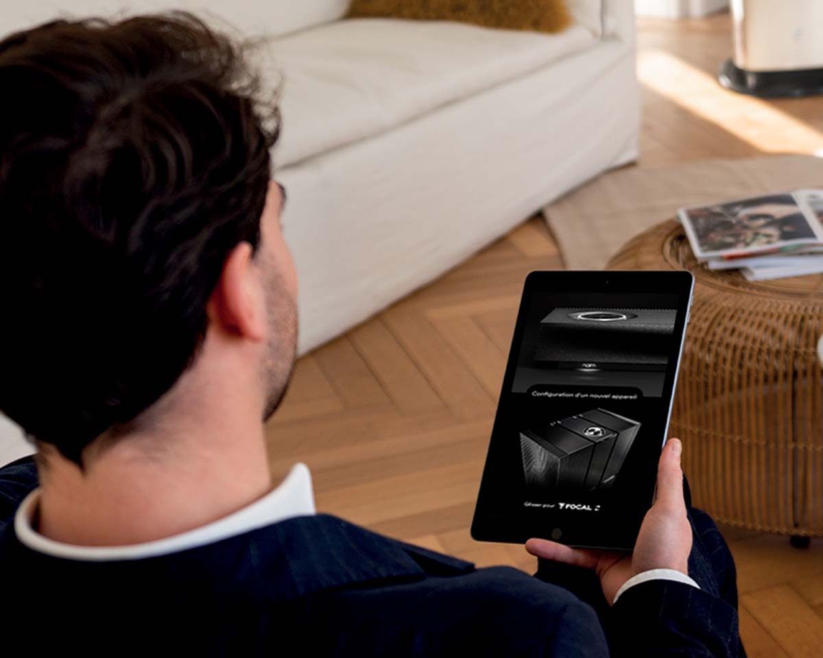 A man in a suit sits on a beige couch, holding a tablet displaying sound equipment settings. A wicker table with magazines is nearby, creating a relaxed atmosphere.