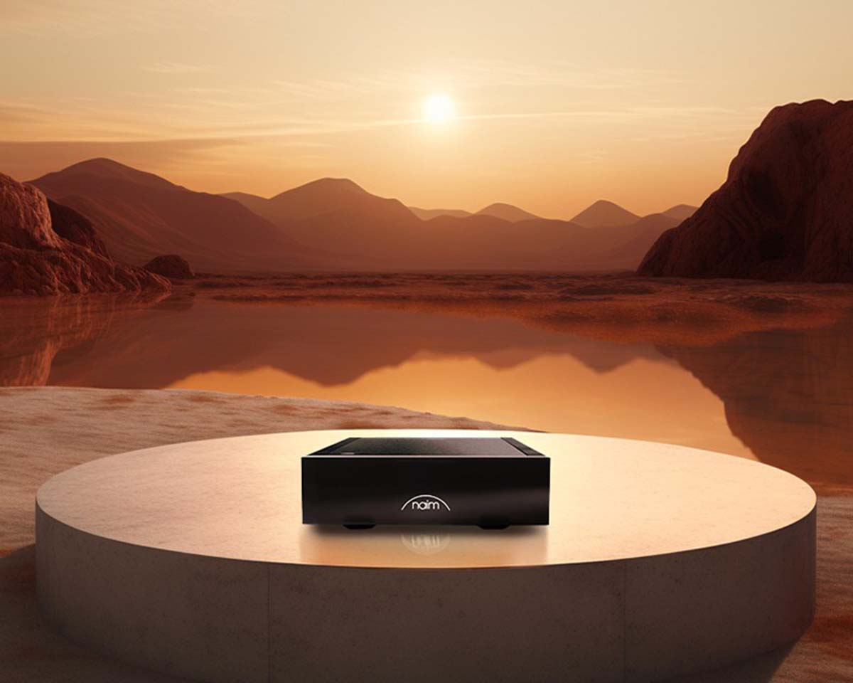 A sleek black audio device sits on a circular platform against a serene sunset backdrop of reddish mountains and a reflective lake, evoking tranquility.
