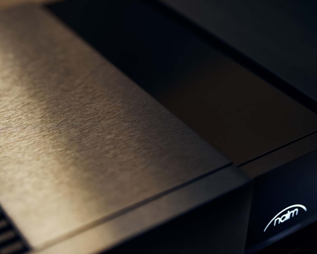 Close-up of a sleek, dark metallic audio device with a brushed finish. A soft glow highlights the brand logo in the lower right corner, giving a modern and sophisticated feel.