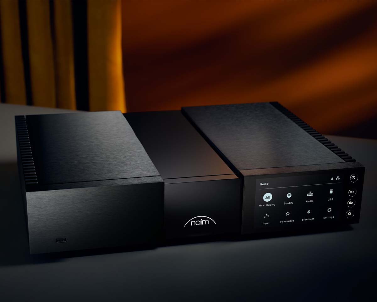 A sleek black audio system with a digital display showing icons like Spotify and radio. "Naim" is illuminated, set against a soft amber background.