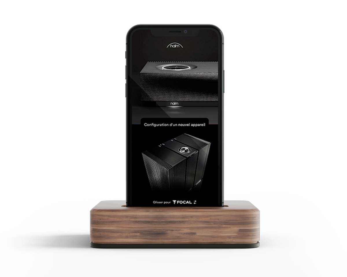 Smartphone on a wooden dock displaying Naim audio setup interface, featuring sleek black speakers with a modern, sophisticated design.