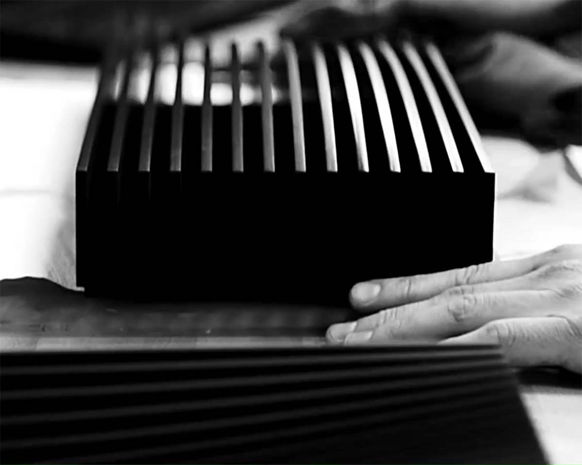 A pair of hands touches a black, finned object, possibly an electronic device, on a table. The monochrome image conveys an industrial, sleek feel.
