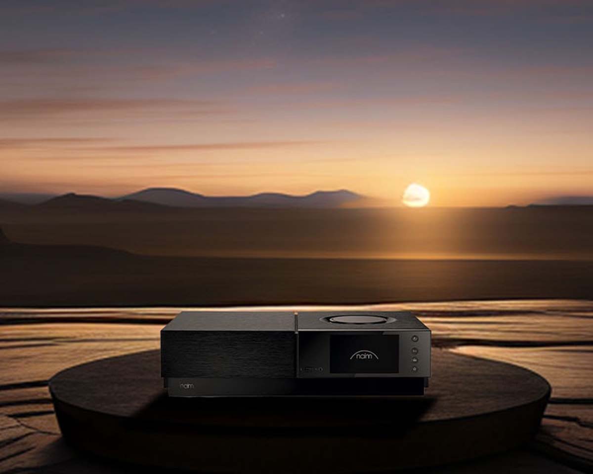 A sleek black audio device sits on a round table against a serene sunset, casting a warm glow over distant mountains. The mood is calm and sophisticated.