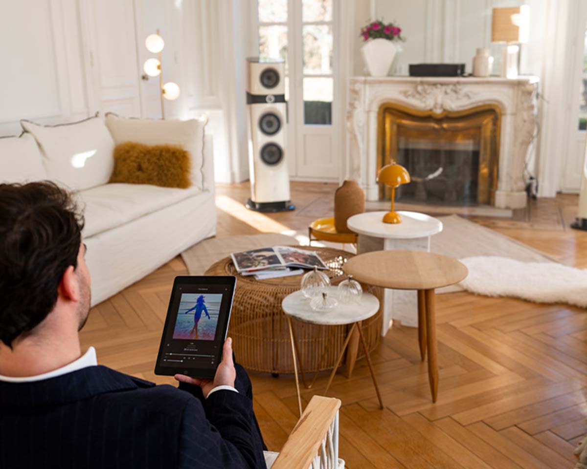 A man seated in a stylish living room holds a tablet displaying music. The room features elegant decor, a fireplace, and large speakers, creating a cozy, high-tech ambiance.