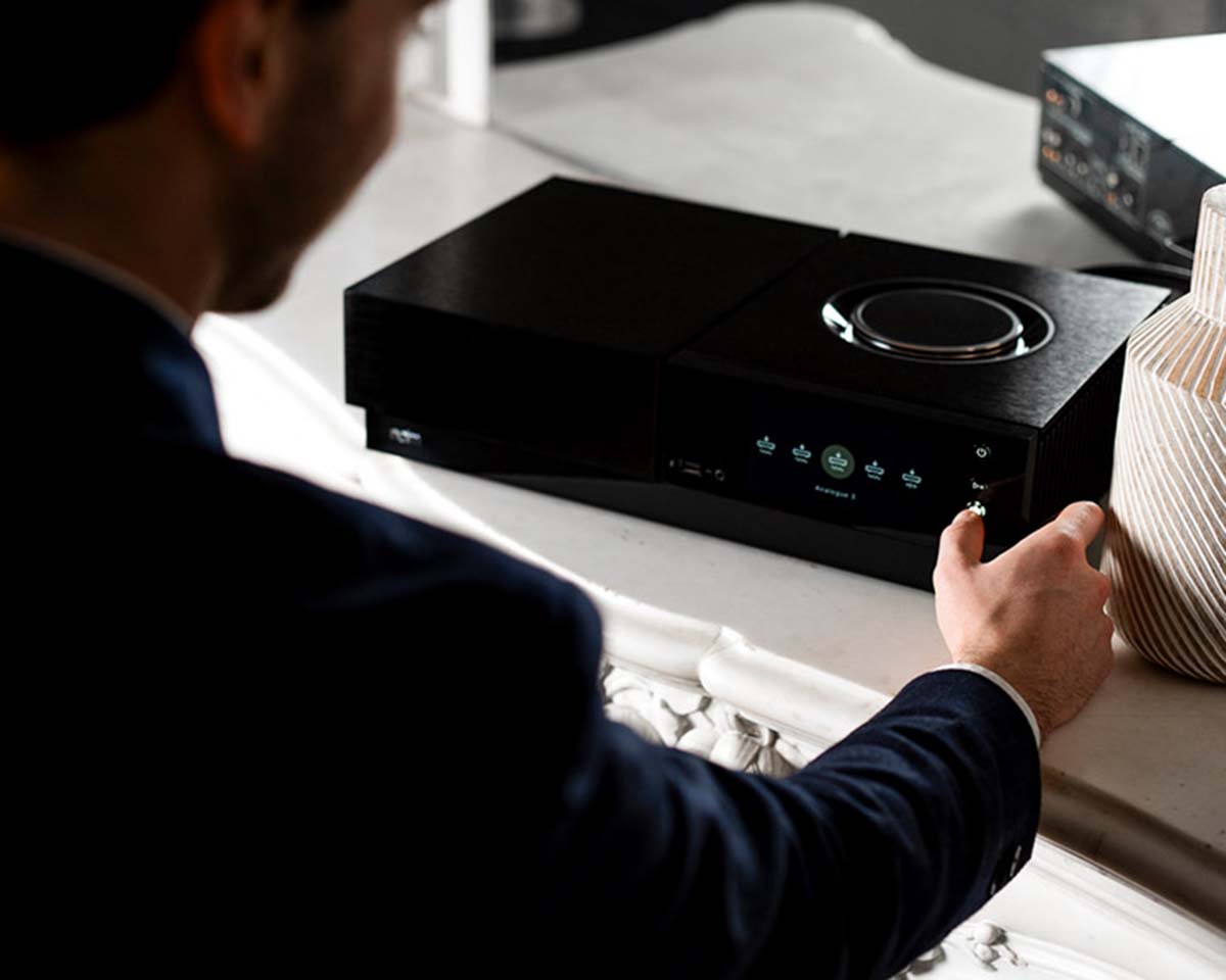A person in a suit adjusts the volume knob on a sleek black audio system with a digital display on a white table, creating a modern, sophisticated ambiance.