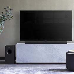 A modern living room features a flat-screen TV on a white marble stand, accompanied by a sleek soundbar and subwoofer. A plant adds a touch of greenery.