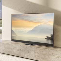 A flat-screen TV displays a serene sunset over a tranquil lake with mountains in the background, set on a simple, modern wooden stand in a minimalist room.
