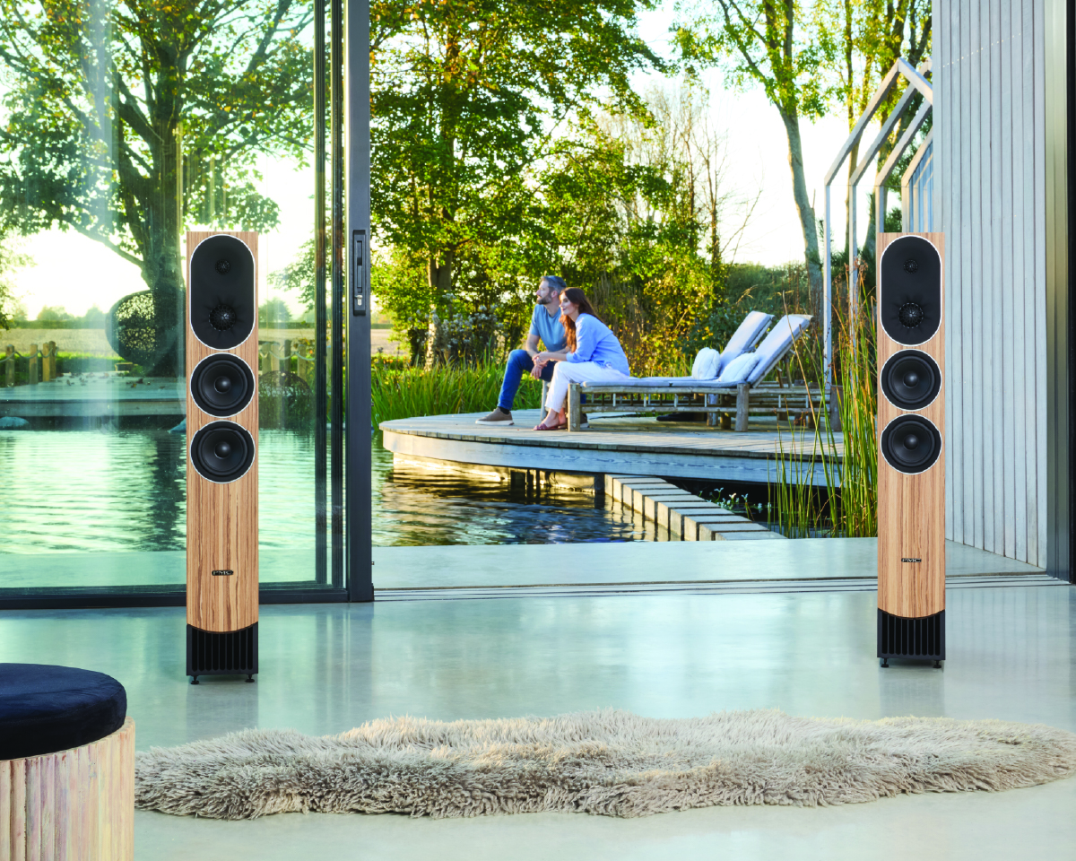 Modern living room with large floor speakers, glass doors open to a serene pond. A couple sits on a deck, surrounded by lush trees. Relaxed ambiance.