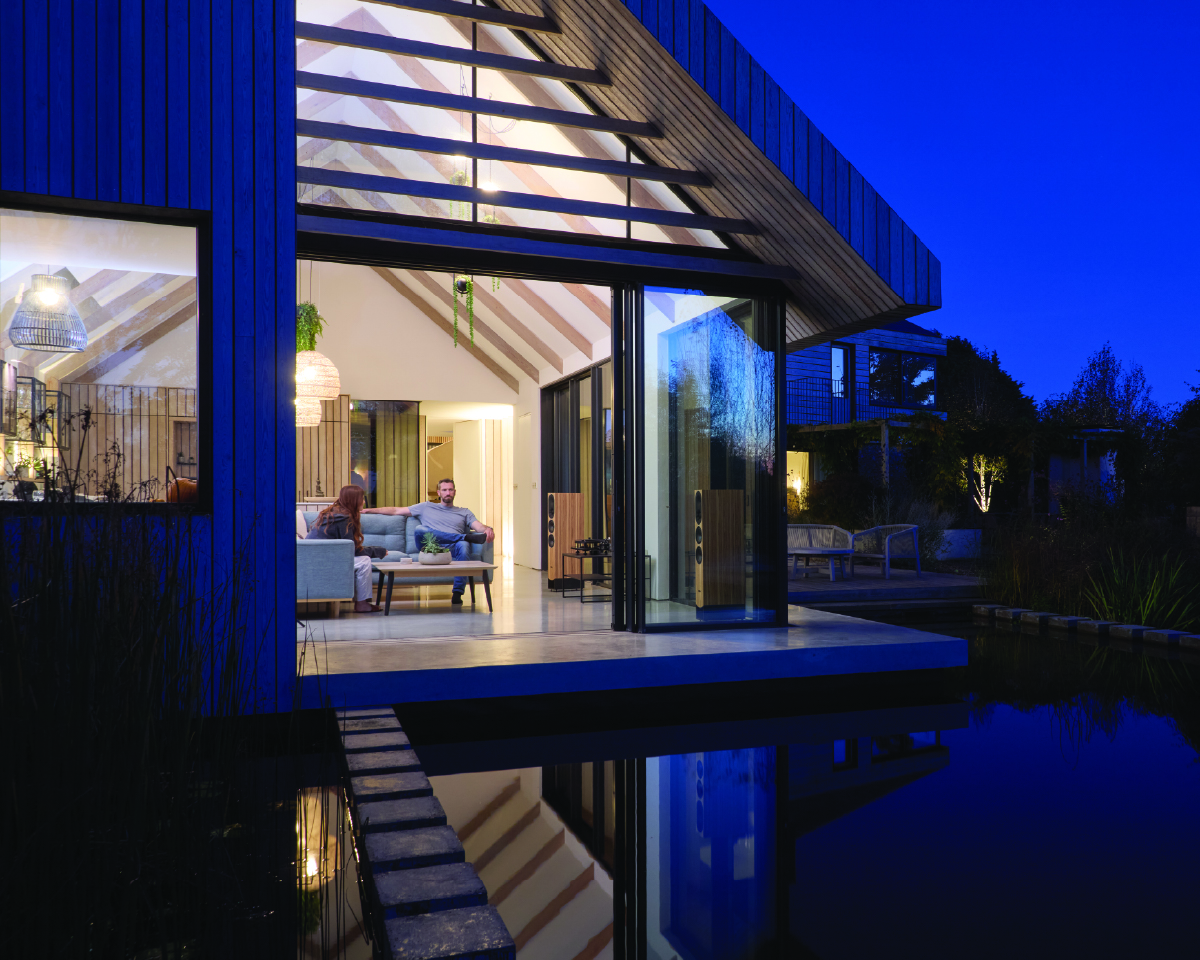 Modern house with large glass walls illuminated at night. Inside, a couple sits on a sofa, creating a cozy and inviting atmosphere. Reflections shimmer on a pond.