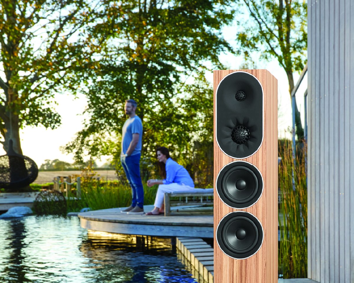 A tall wooden speaker stands prominently by a tranquil pondside deck. In the background, a man stands and a woman sits, both in a relaxed outdoor setting.
