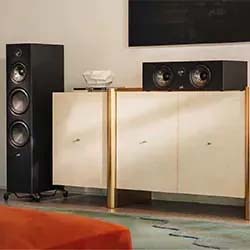 A sleek, modern living room with a tall black speaker and a cabinet topped with a center speaker and decorative bowl on a patterned rug with a cozy orange corner.