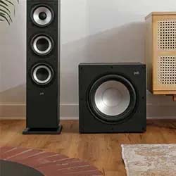 A black floor-standing speaker and a subwoofer on a wooden floor, near a beige cabinet. The setup conveys a modern and minimalistic home audio system.