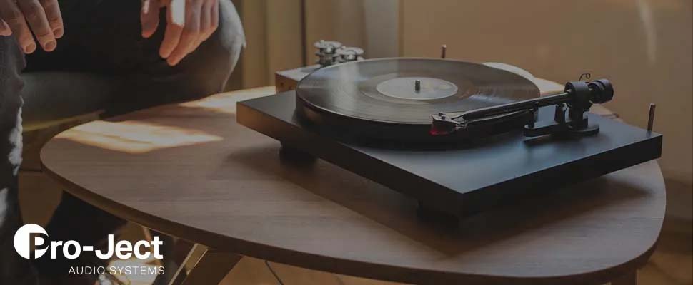 A vinyl record player sits on a wooden table in a sunlit room. A person seated nearby reaches towards it, creating a cozy, nostalgic atmosphere. Logo in corner.