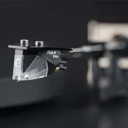 Close-up of a turntable stylus labeled "Pick It Pro," with a sleek black and silver design against a blurred dark background, conveying a modern, high-tech feel.