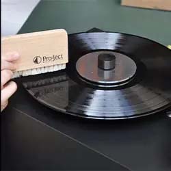 A hand holds a brush with wooden handle cleaning a black vinyl record on a turntable. The setting is minimal, conveying a sense of care and nostalgia.
