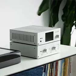 A modern stereo system with silver components, featuring dials and a screen, sits on a white shelf next to books and a green plant, creating a sleek, contemporary vibe.