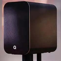Sleek black speaker with rounded edges on a stand, highlighted by soft lighting. The minimalist design, featuring a white "Q" logo, conveys modern elegance.