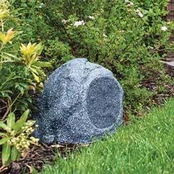 A rock-shaped speaker is nestled among lush green plants and grass, blending seamlessly into a garden setting. The scene conveys a natural ambiance.