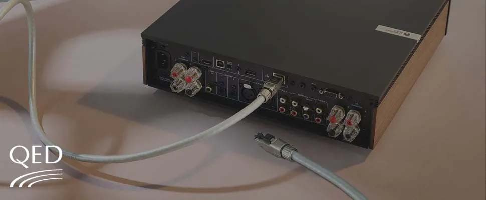 Back view of a black audio amplifier featuring multiple input and output ports, with cables connected. The setting is neutral and technical.